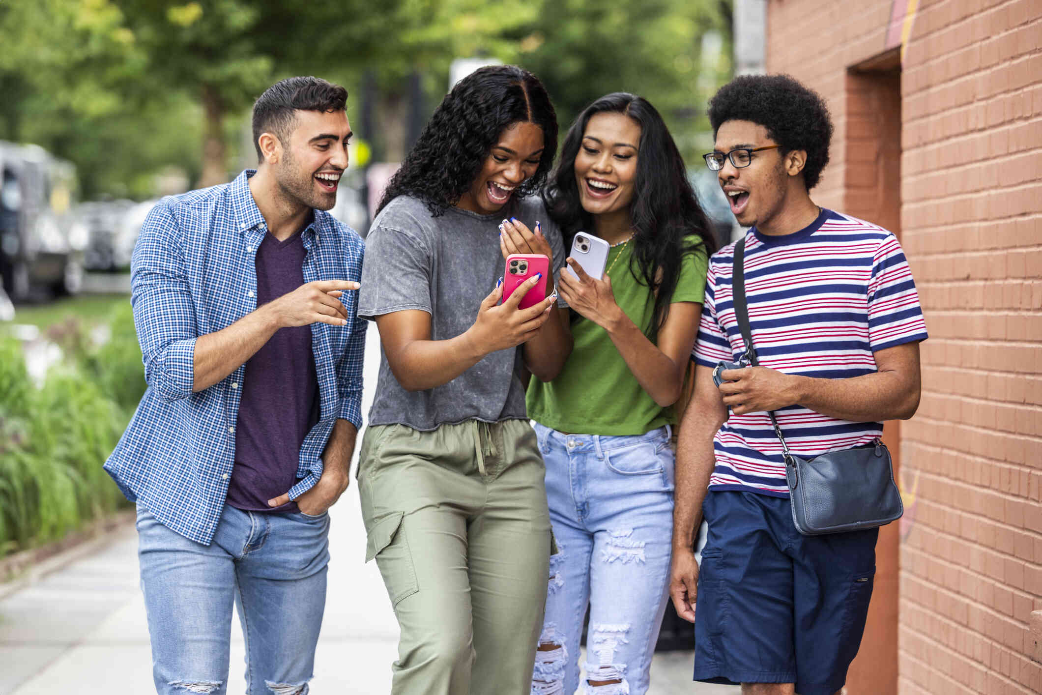 A group of adult friends walk together outside on a sunny day while looking at the phone in one of the girls hands while smiling and laughing.